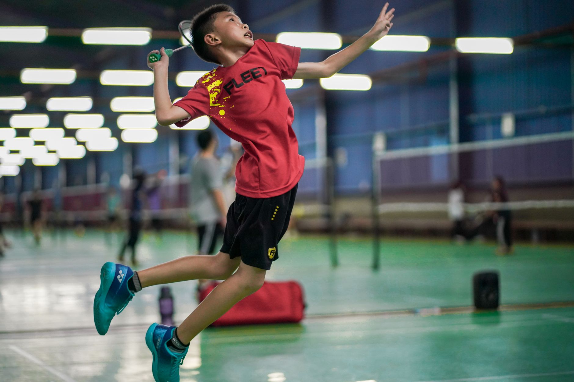 A young boy enthusiastically jumps to hit a badminton shuttlecock inside a sports hall