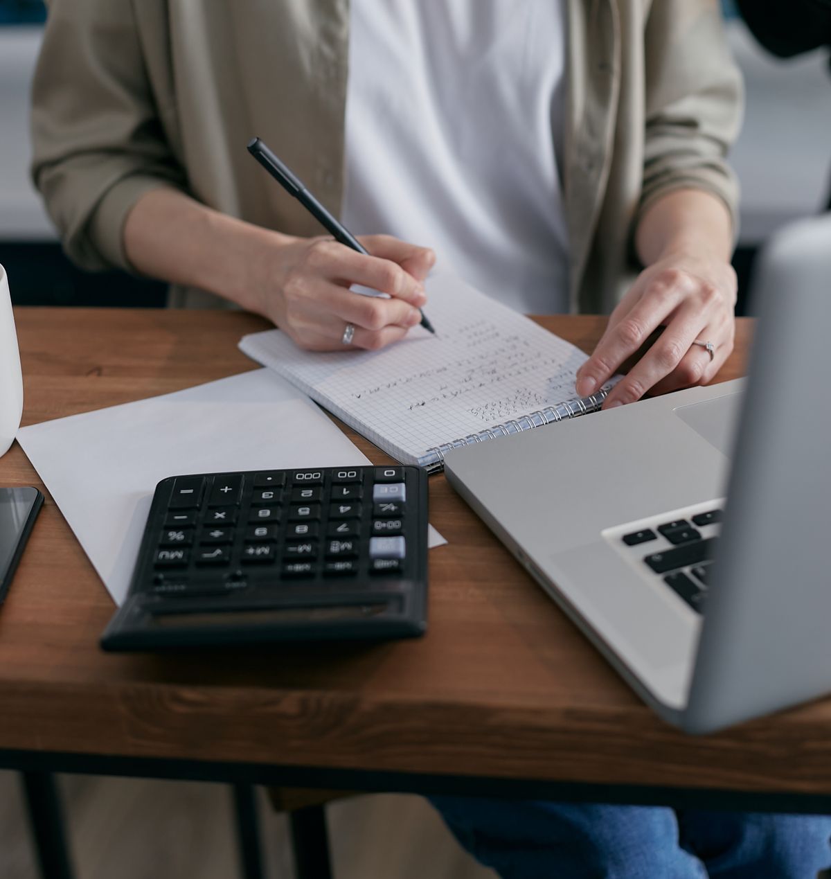 Person calculating figures in a notebook beside a laptop on a desk