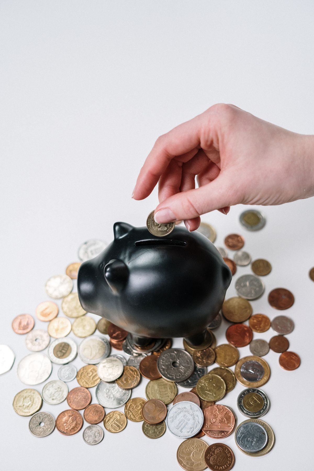 Hand placing a coin into a black piggy bank surrounded by coins