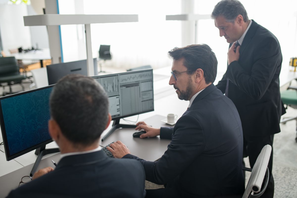 Three men in business suits gathered around computer screens in a professional office
