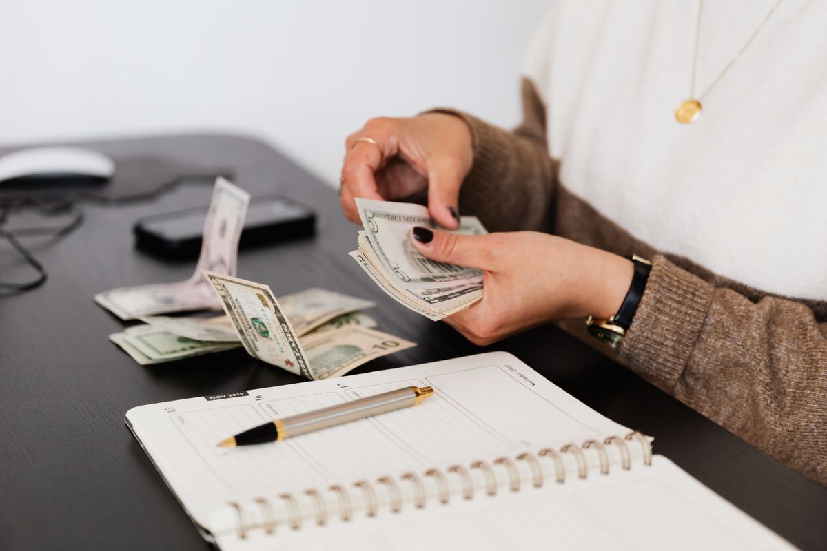 A person counting a stack of banknotes beside a notepad on a desk