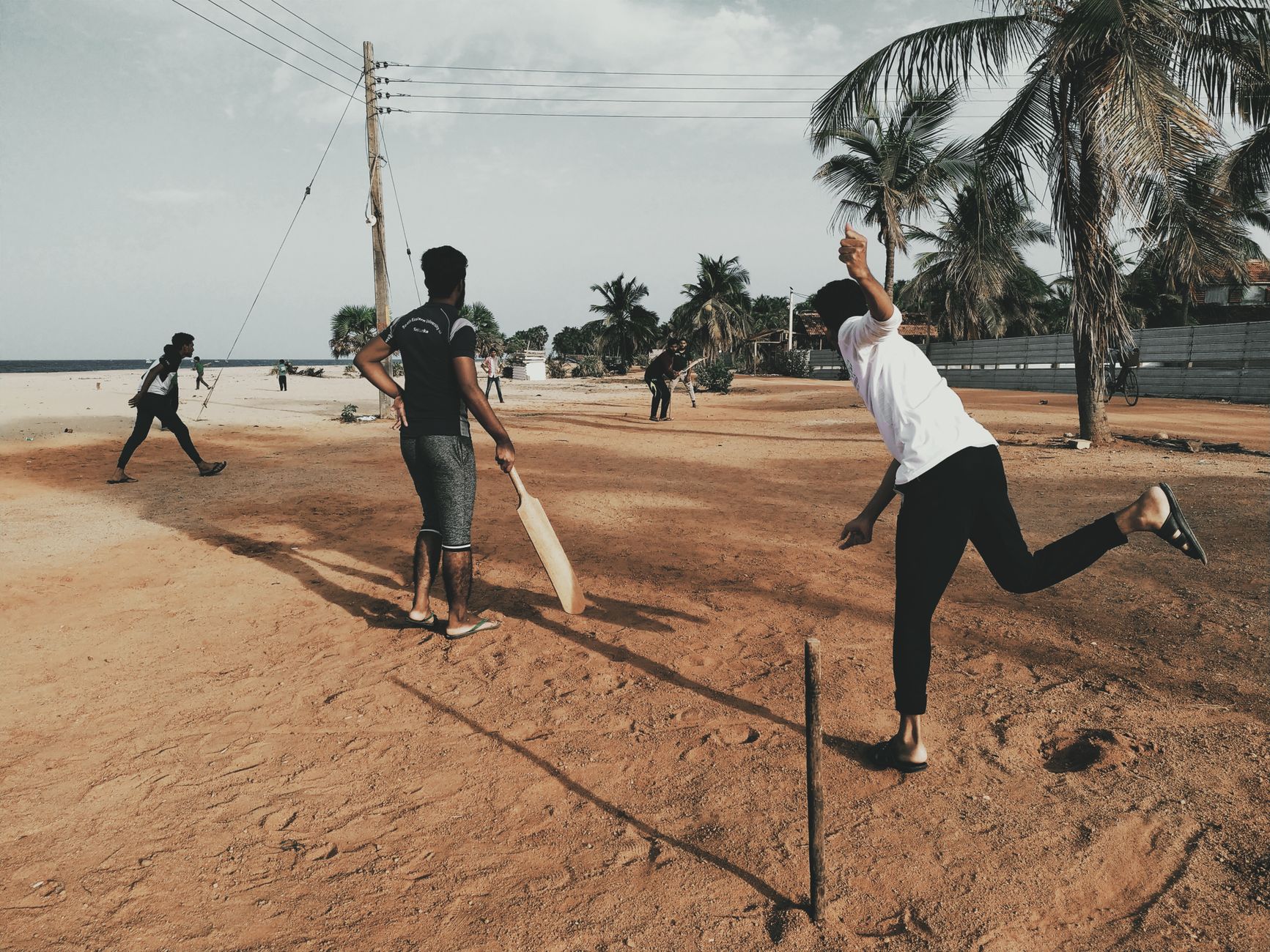 A group of young men playing cricket on a sandy beach under clear skies