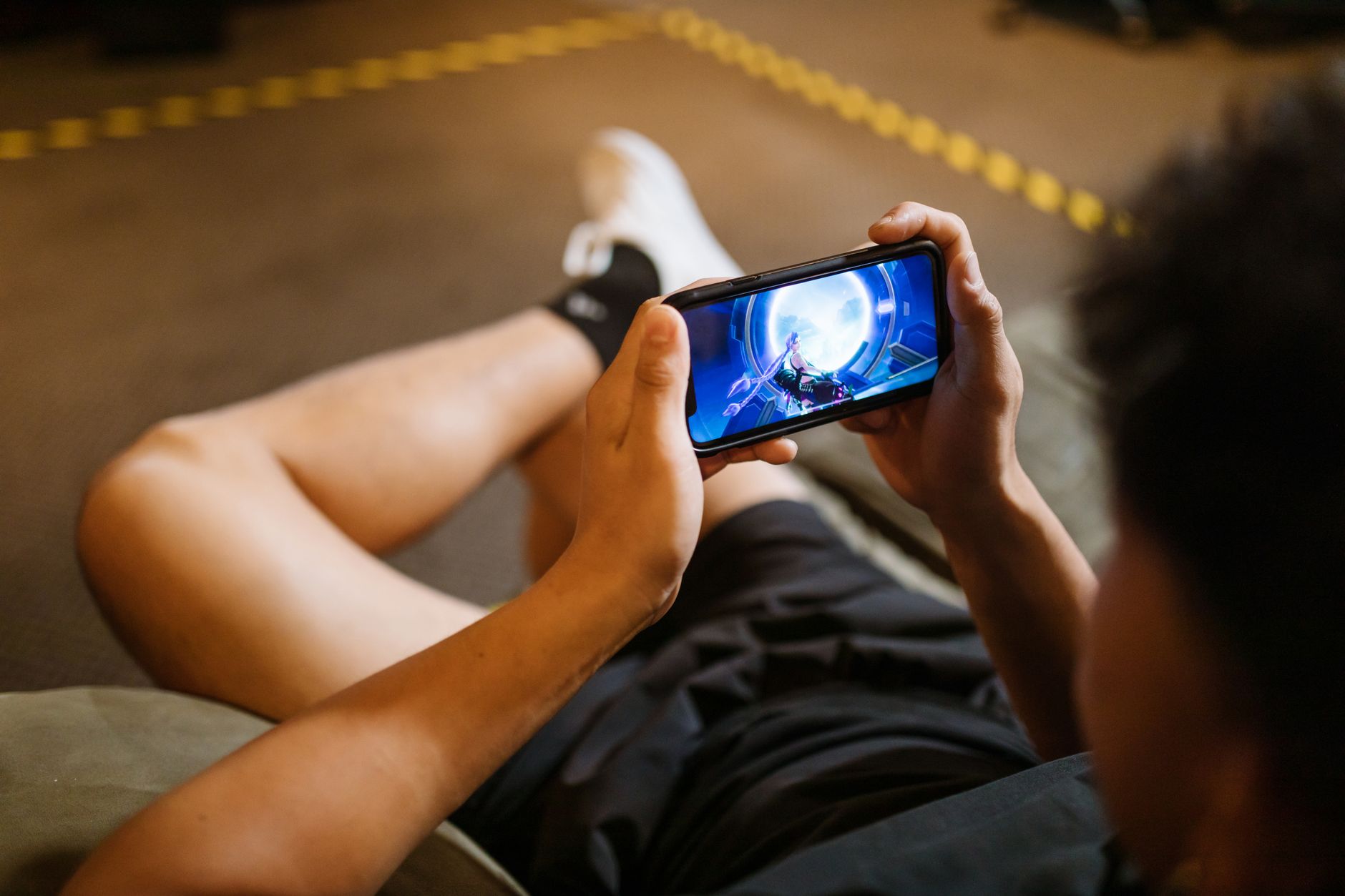 Young players in a dim room lit by a phone screen, one holding a mobile phone playing a battle royale game