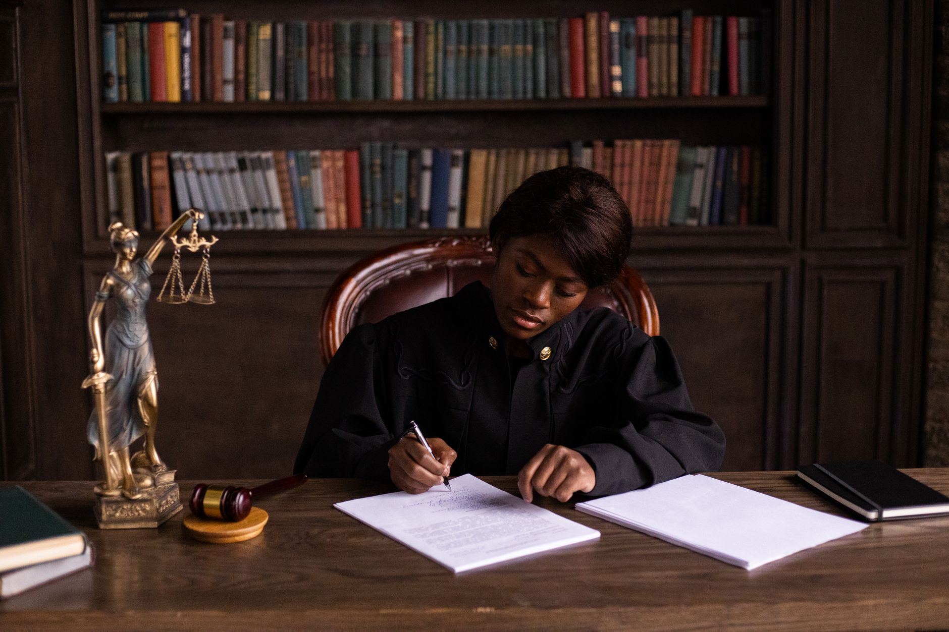 A judge in robes writes on a document at a desk with scales of justice and law books behind her