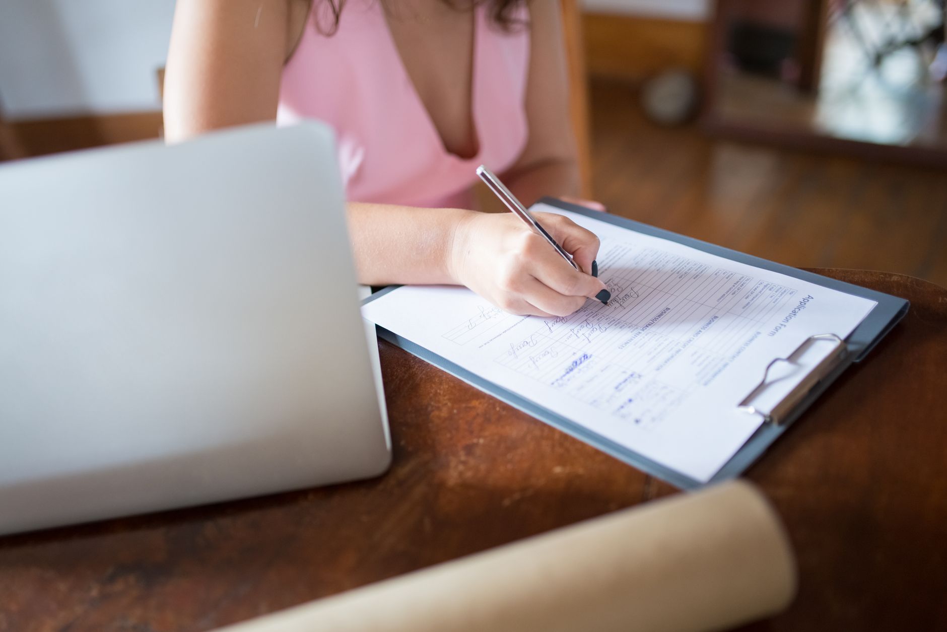 A person fills in a form on a clipboard beside a laptop on a wooden desk
