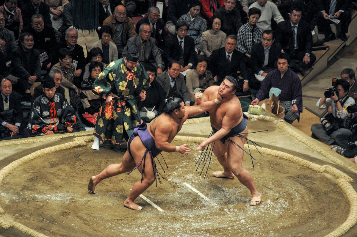 Two sumo wrestlers grappling at the tachiai inside a clay dohyo ring in a Japanese arena
